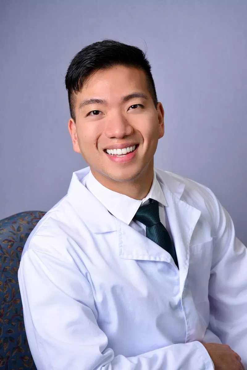 Professional male dentist in white coat and tie smiling at camera in clinical setting.