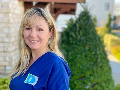 Smiling dental professional in blue scrubs standing outside brick building with evergreen landscaping.