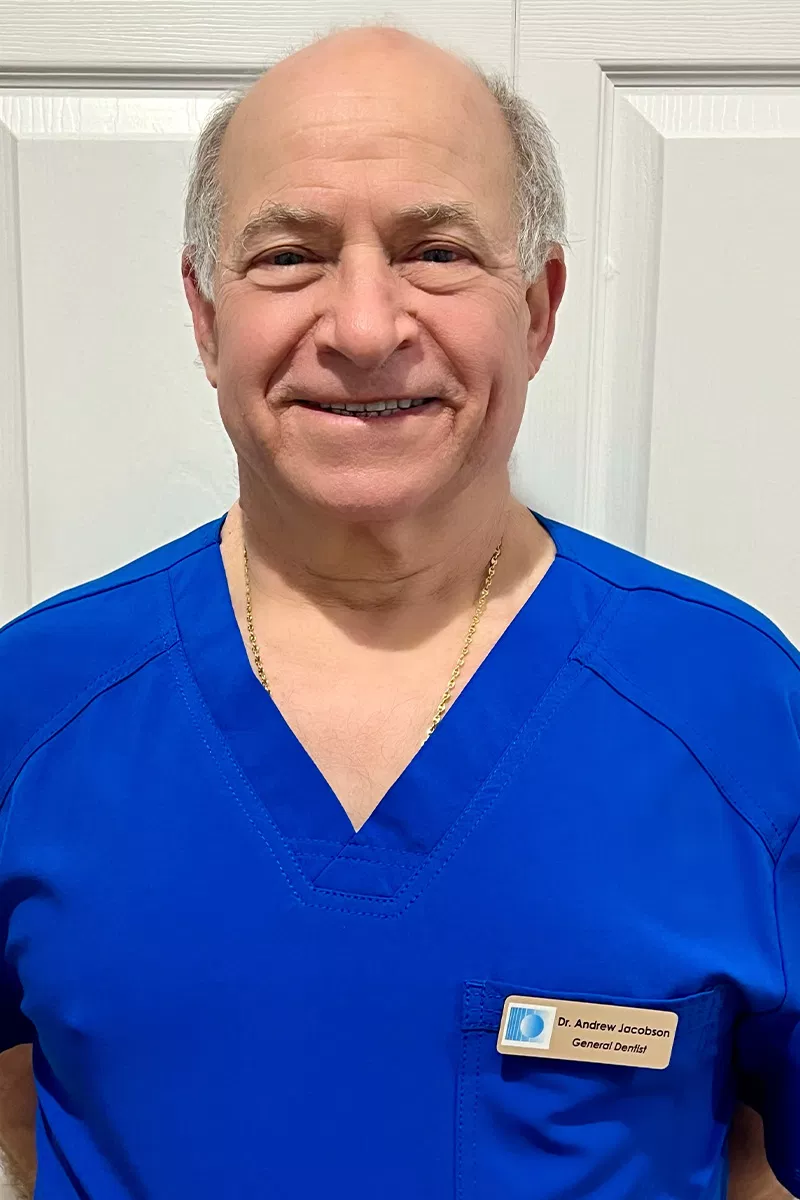 Male dentist in blue scrubs smiling at camera in dental office setting.