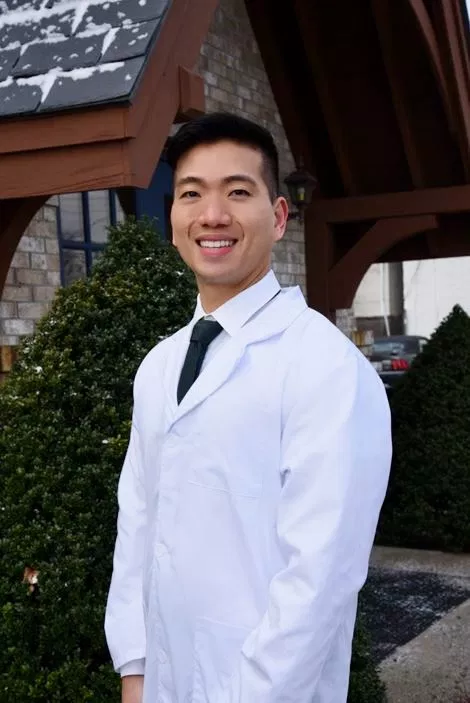 Male dentist in white coat smiling outside dental office building with landscaping.