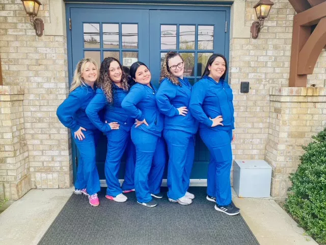 Dental team of six women in blue scrubs posing together outside brick building entrance.
