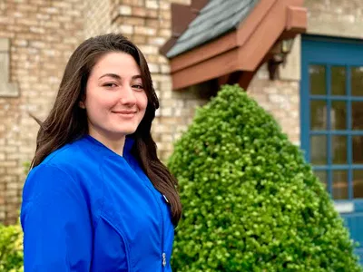 Woman in blue scrubs smiling outside brick dental office building with ivy.