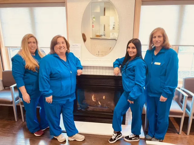 Four dental staff members in blue scrubs pose together in a modern dental office waiting room with fireplace.
