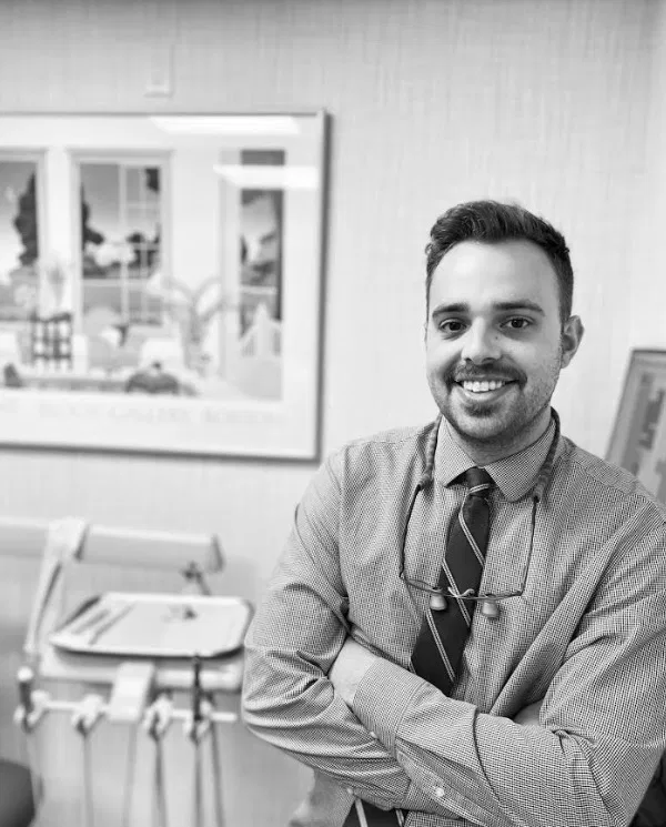 Male dentist smiling at desk in modern dental office with equipment visible.