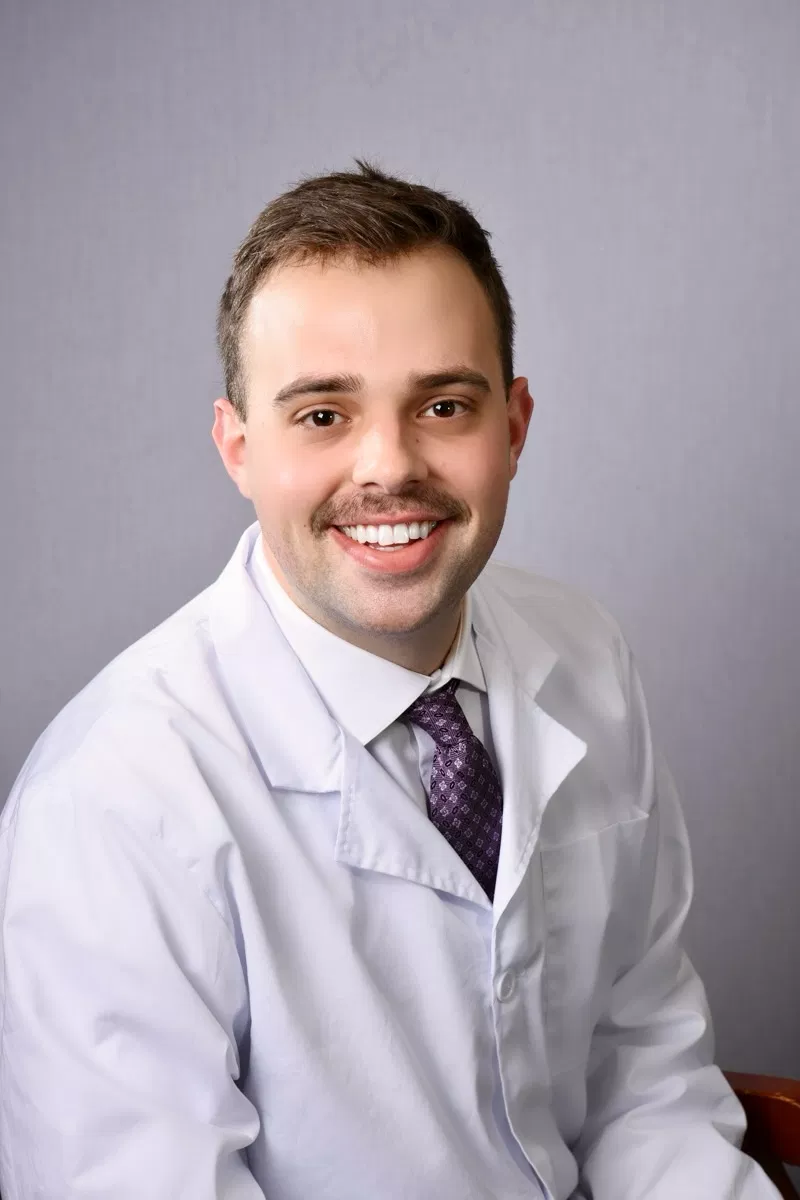 Professional male dentist in white coat and purple tie smiling at camera against gray background.