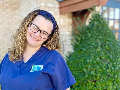 Smiling dental professional in blue scrubs wearing glasses outside brick building with ivy.