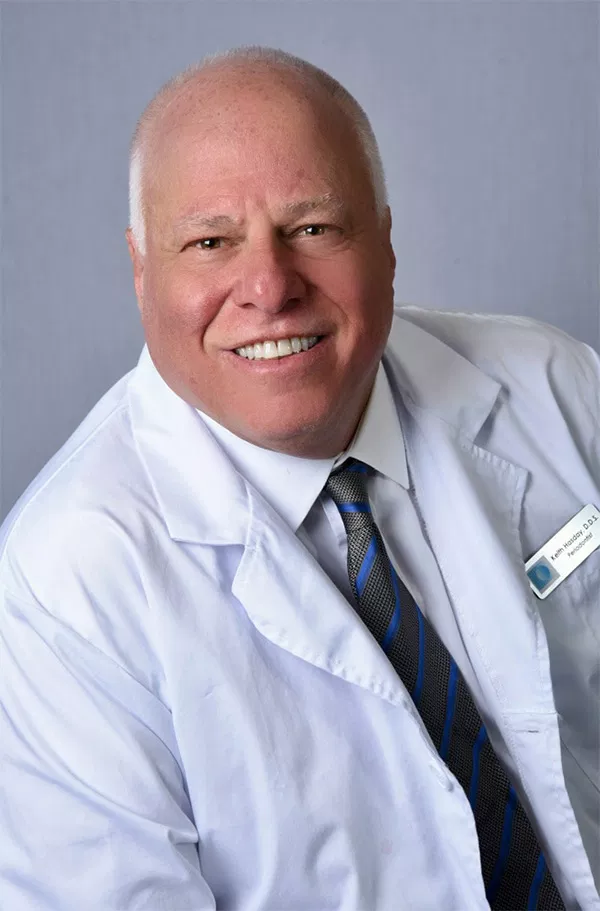 Professional male dentist in white coat and tie smiling at camera in clinical setting.