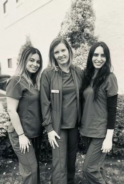 Three female dental professionals in scrubs posing together outdoors near evergreen shrub.