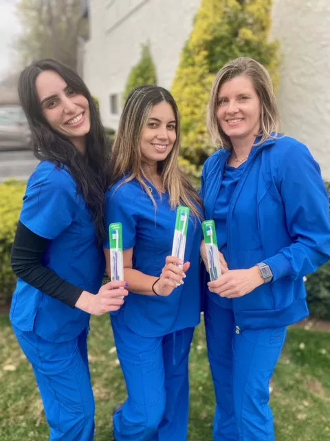 Three women in blue scrubs holding certificates outside a dental office building.