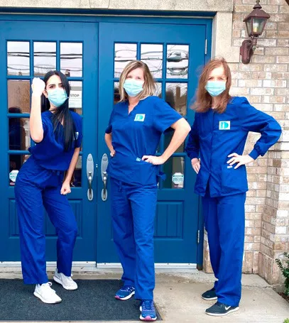Three dental professionals in blue scrubs and masks standing outside blue entrance doors.