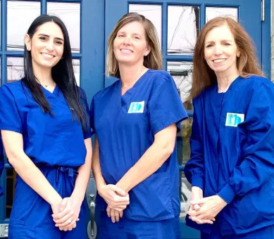 Three female dental professionals in blue scrubs standing together outside a dental office entrance.