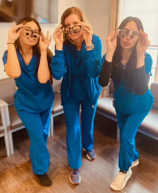 Three dental professionals in blue scrubs adjusting glasses and smiling in a clinical office setting.