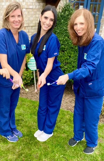 Three dental professionals in blue scrubs standing together outdoors on grass.