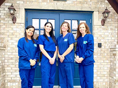Four female dental professionals in blue scrubs posing together in front of brick building with blue doors.