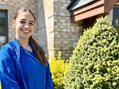 Smiling dental professional in blue scrubs stands outside modern brick dental office building.