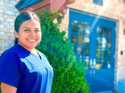 Smiling female dental professional in blue scrubs standing outside modern blue dental office building.