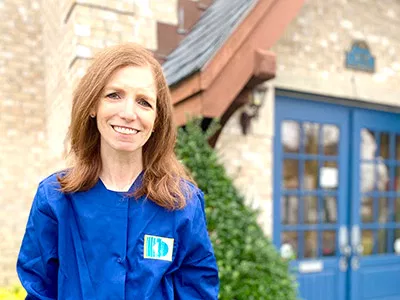 Woman in blue dental scrubs smiling outside brick building with blue doors.