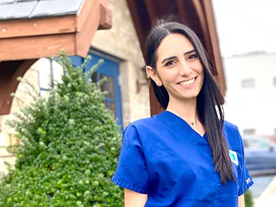 Female dental professional in blue scrubs smiling outside modern dental office building.