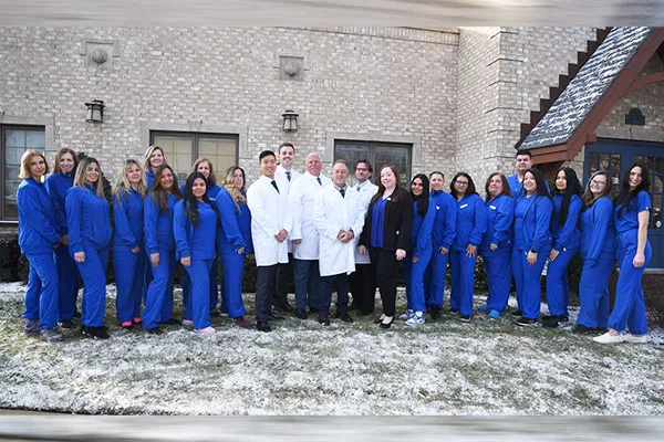 Dental team staff in blue scrubs and white coats posing together outside brick building.