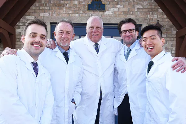 Five male dentists in white coats posing together in a dental office with brick wall backdrop.