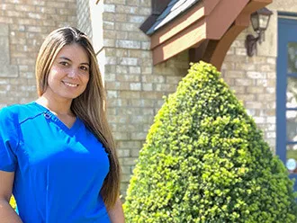Woman in blue scrubs smiling outside brick dental office building with green topiary.