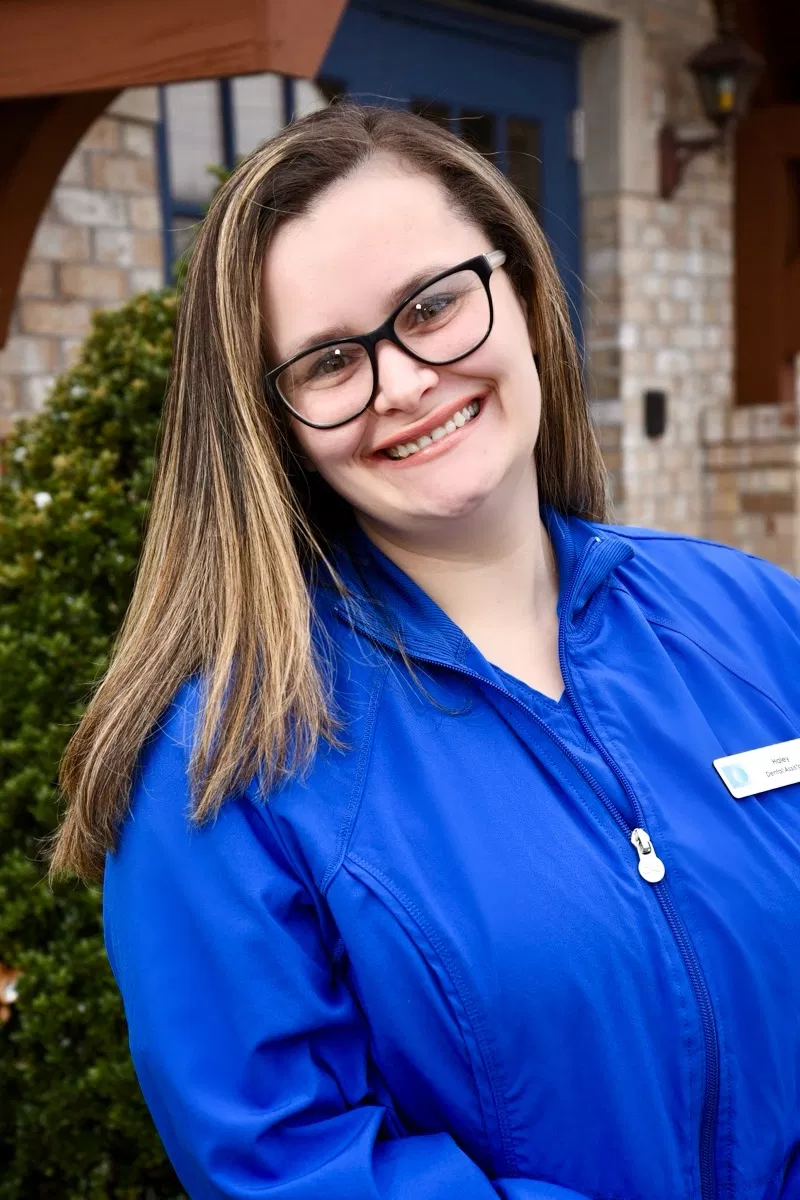 Young woman wearing blue dental scrubs and glasses smiling outdoors near brick building.