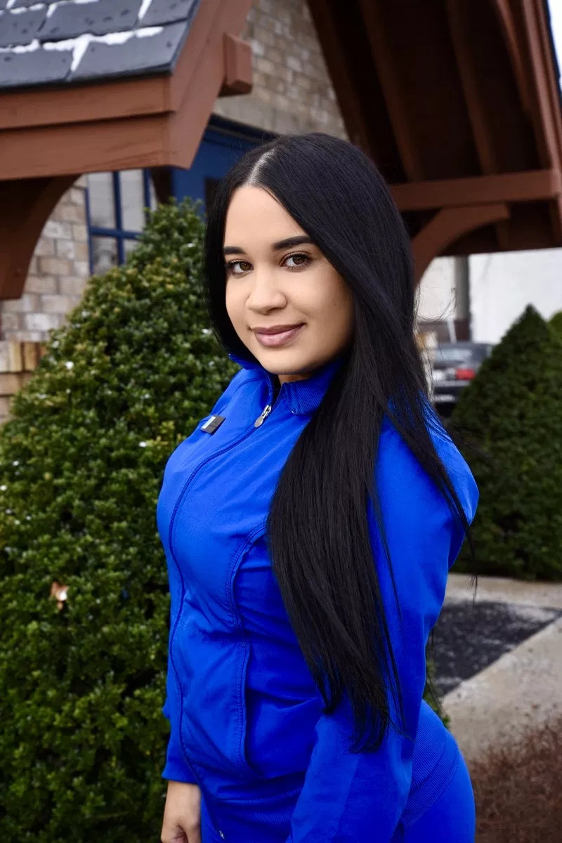 Woman in blue outfit smiling outdoors near building with hedges, professional portrait setting.