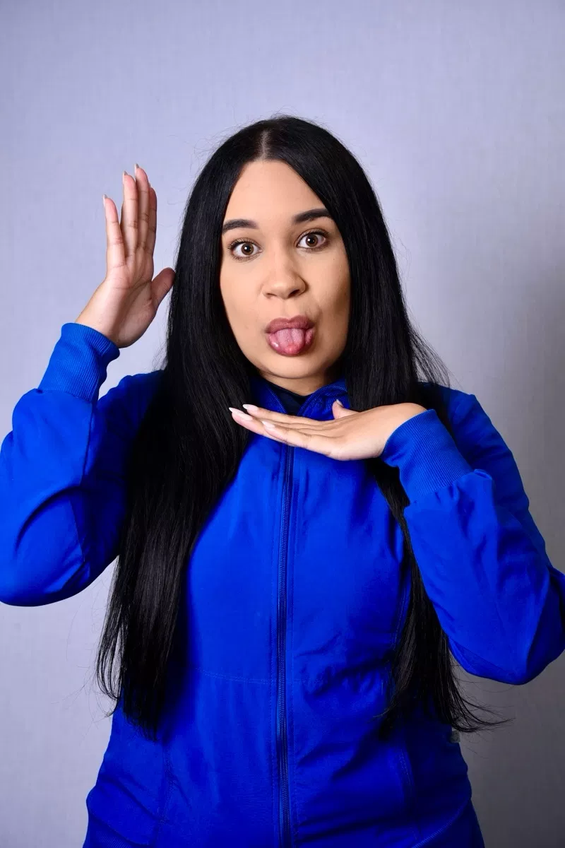 Woman in blue top with surprised expression against gray background, hand near face.