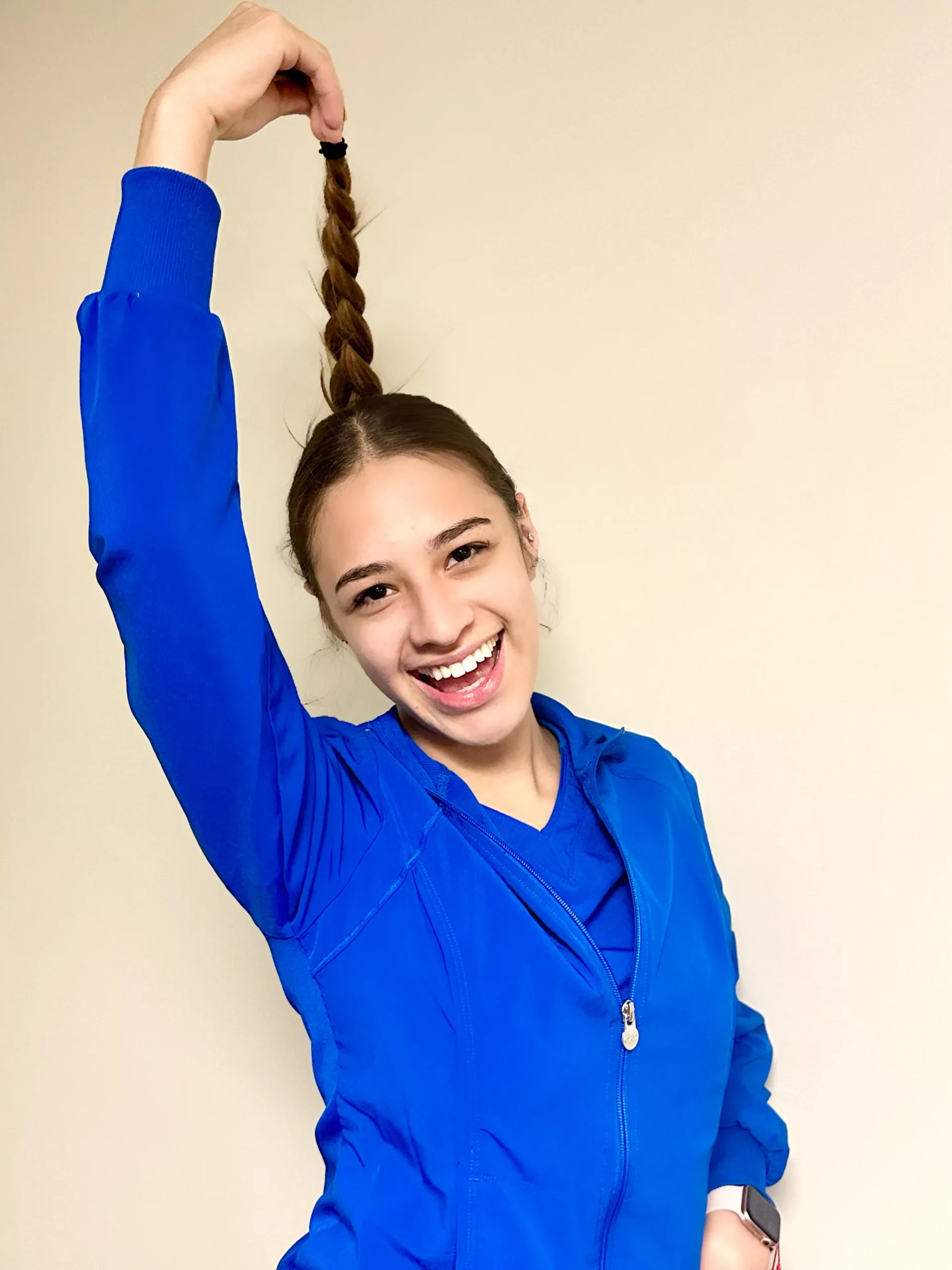 Young girl in blue hoodie holding braided hair up, smiling against cream background.