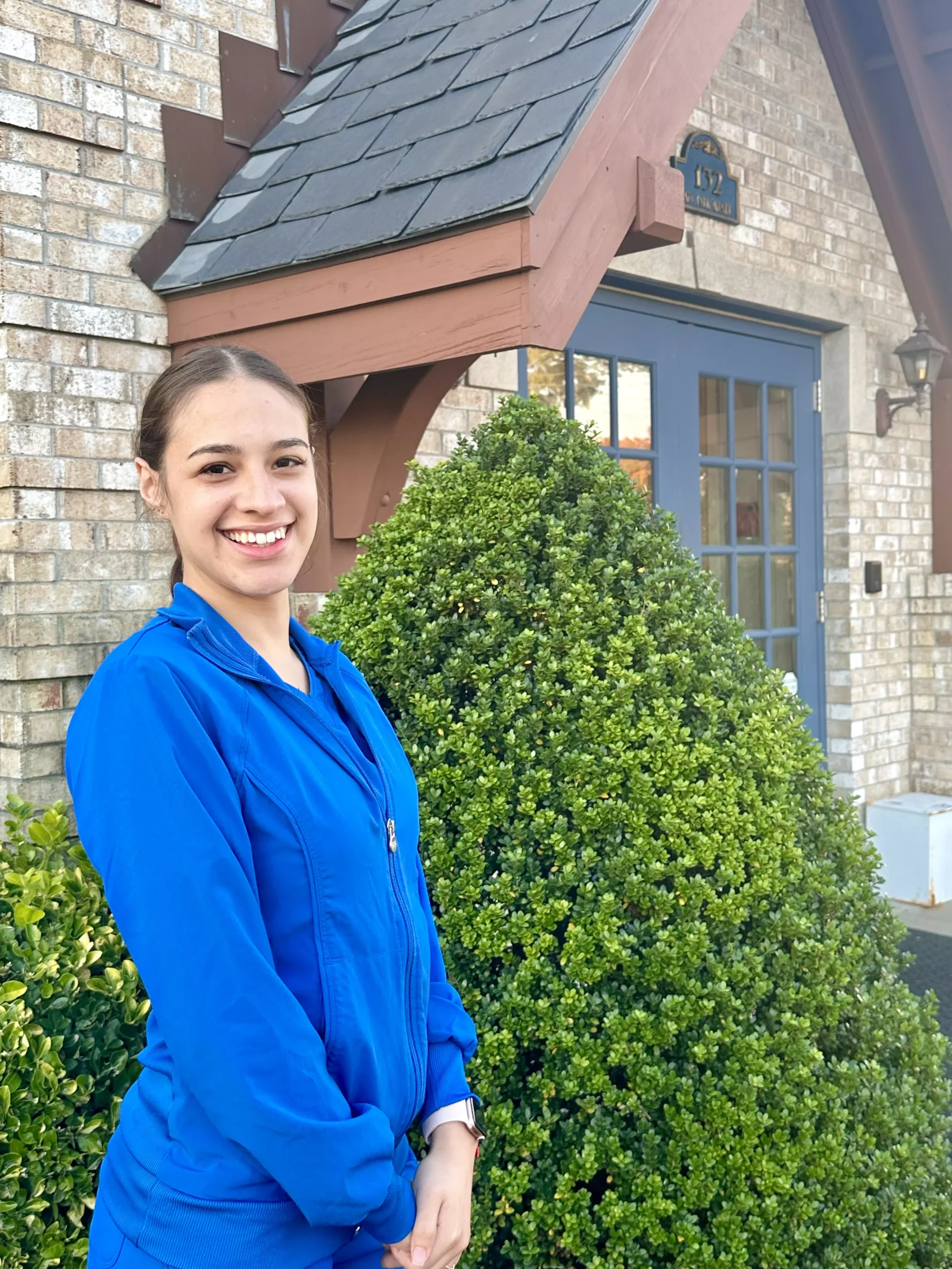 Woman in blue scrubs smiling outside brick dental office building with ivy plants.