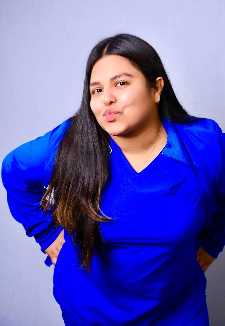 Female dental professional in blue scrubs posing against light purple background for portrait.