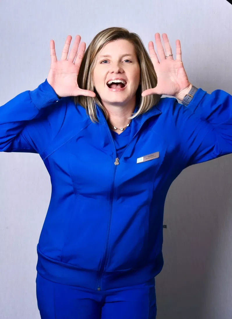 Woman in blue scrubs gesturing excitedly with hands raised near face, dental office setting.