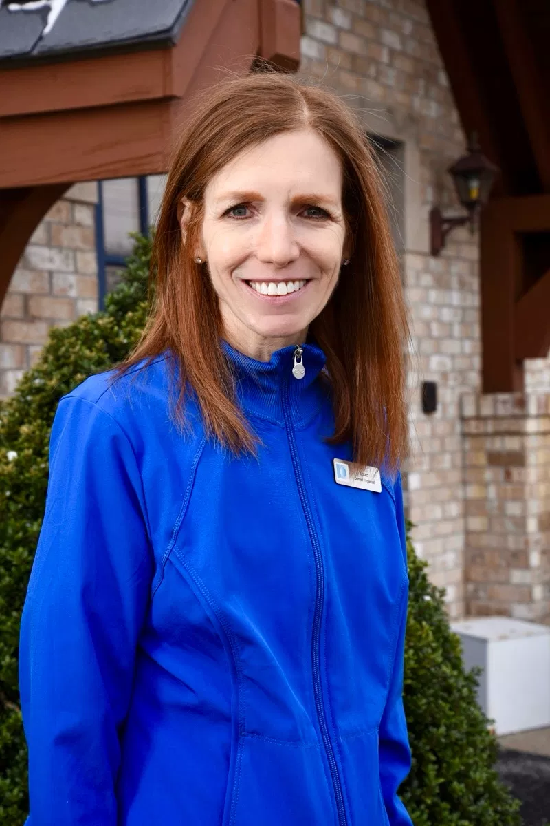 Woman in blue jacket smiling outdoors in front of brick building entrance.