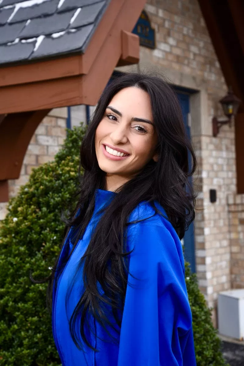Young woman with long dark hair smiling in blue jacket outside brick building.