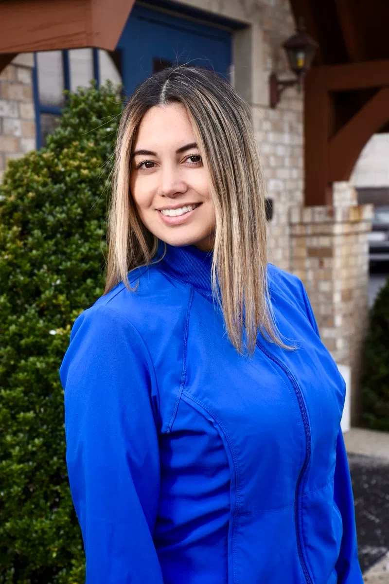 Professional woman in blue shirt smiling outdoors at dental office entrance with brick building.