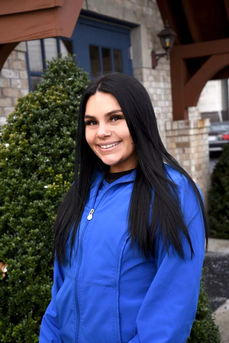 Woman in blue jacket smiling outdoors near ivy-covered building entrance.