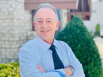 Professional male dentist in light blue shirt and tie, arms crossed, smiling outdoors.