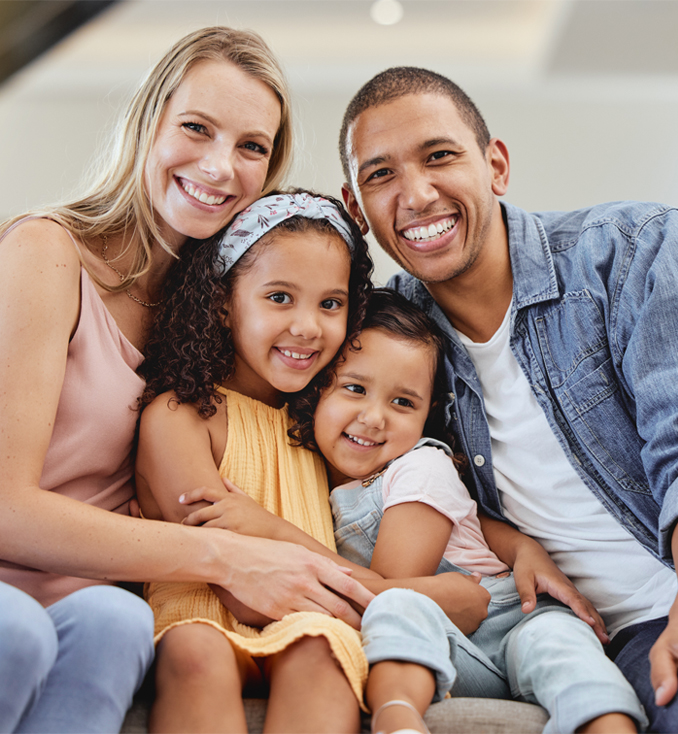 Happy multiracial family of four smiling together indoors, embracing in casual clothing.