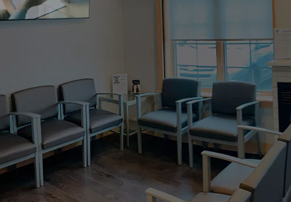 Modern dental waiting room with gray upholstered chairs and side table near windows.