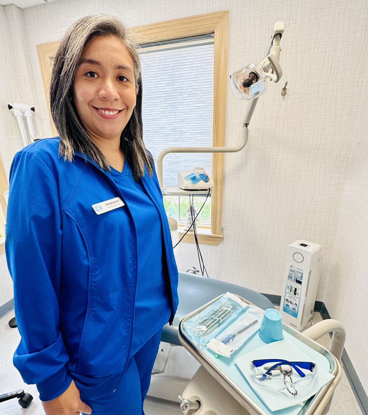 Dental hygienist in blue scrubs smiling in modern treatment room with equipment.