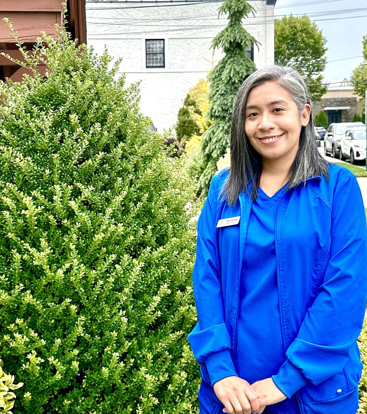 Woman in blue dental scrubs smiling outdoors next to green shrubs.