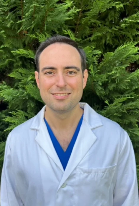 Male dentist in light blue coat and blue scrubs smiling outdoors with green foliage background.