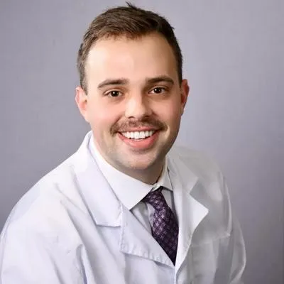 Professional male dentist in white coat and tie smiling at camera in clinical setting.