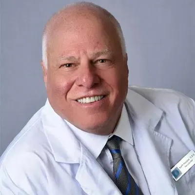 Male dentist in white coat and blue tie smiling at camera in professional portrait.