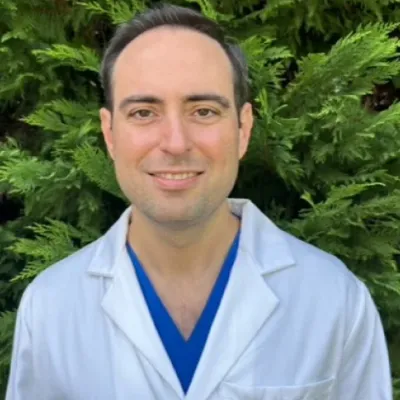 Male dentist in white coat and blue scrubs smiling outdoors with green foliage background.