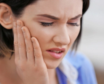 Woman holding jaw in pain, appearing to experience tooth or dental discomfort indoors.