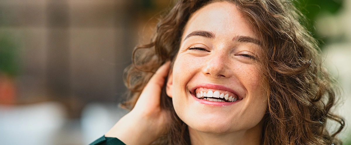 Smiling woman with curly hair touching her face in bright outdoor setting, promoting dental health and wellness.