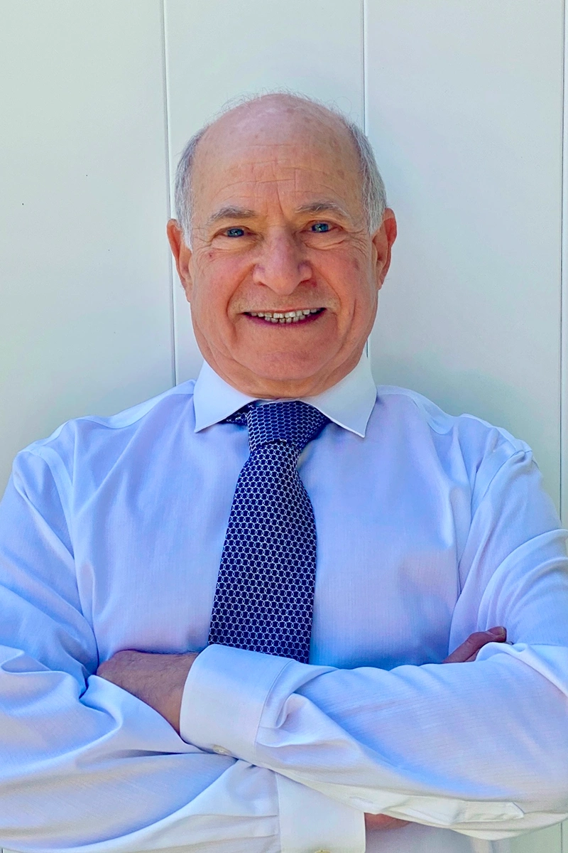 Professional dentist in blue shirt and tie, smiling at camera in clinical office setting.