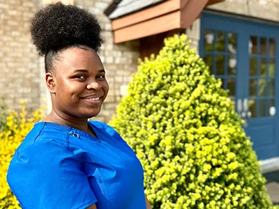 Woman in blue shirt smiling outdoors near bright green shrub and building entrance.