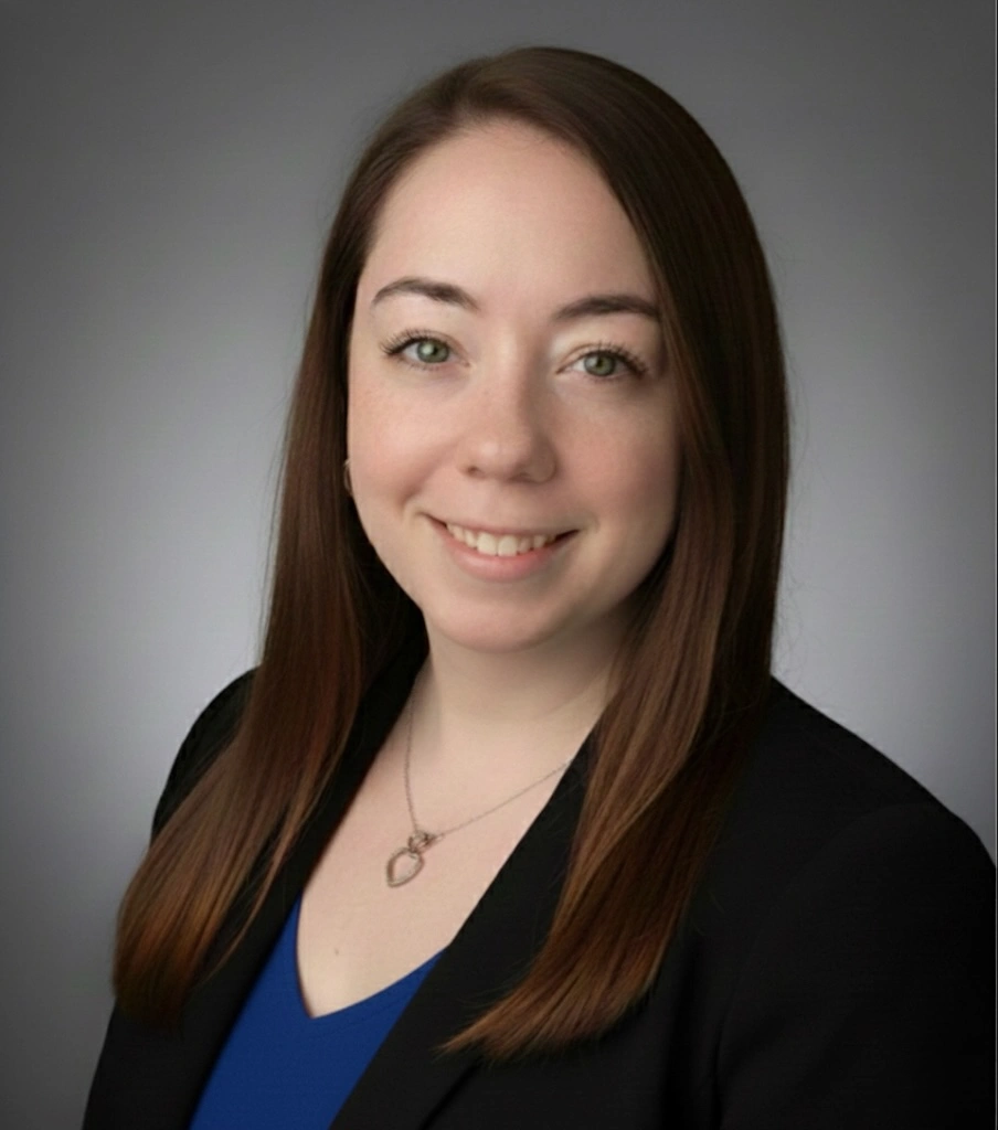Professional headshot of smiling woman in blue top and black blazer against gray background.